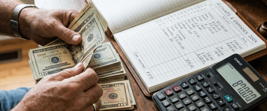 Person counting cash beside an Accounts & Expenses 2024 ledger and calculator